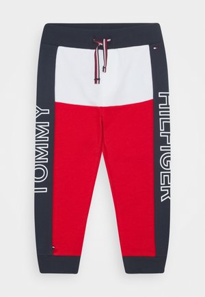Pantalon de survêtement rouge et blanc avec une bande latérale bleu marine portant le texte "TOMMY HILFIGER". Taille avec cordon de serrage et rayures contrastées. Tissu doux au toucher.