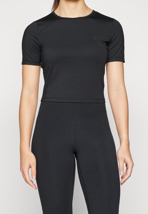 Kobieta w dopasowanym czarnym krótkim topie z krótkim rękawem i pasujących legginsach z wysokim stanem, stojąca na jednolitym tle.