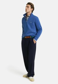 Blauer Fleece-Pullover mit hohem Kragen und Reißverschluss, kombiniert mit dunklen Hosen und hellen Schuhen. Verfügt über eine Tasche und einen braunen Gürtelakzent.