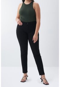 Femme portant un haut sans manches vert foncé, un pantalon noir ajusté et des sandales à talons noires à brides, debout devant un fond uni.