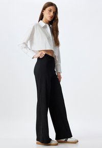 Witte cropped button-up shirt met lange mouwen, gecombineerd met hoge, zwarte wijde broek. Model draagt witte sneakers.