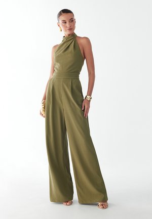 Femme portant une combinaison sans manches vert olive à col bénitier avec jambes larges, bracelets dorés, boucles d'oreilles et sandales, sur un fond blanc uni.