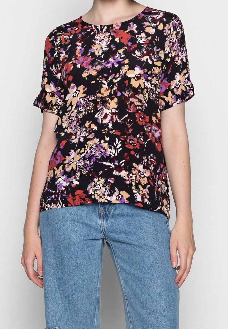 Femme portant un haut noir à manches courtes avec des motifs floraux multicolores et un jean en denim bleu clair, debout sur un fond uni.