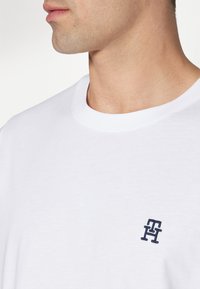 T-shirt bianco in cotone con scollatura a girocollo, caratterizzato da un piccolo logo ricamato blu navy sul lato sinistro del petto. Texture morbida e design semplice.