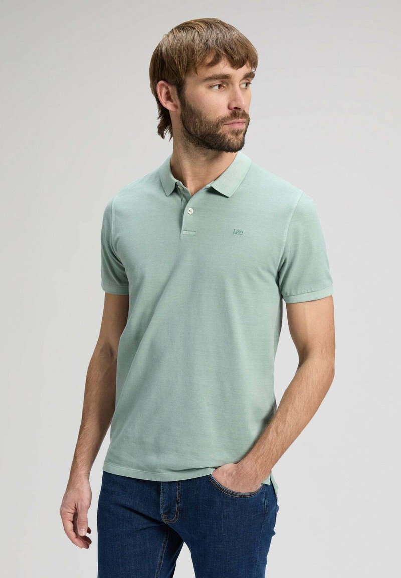 Homme barbu portant un polo vert clair et un jean bleu, debout avec une main dans la poche, regardant sur le côté sur un fond uni.