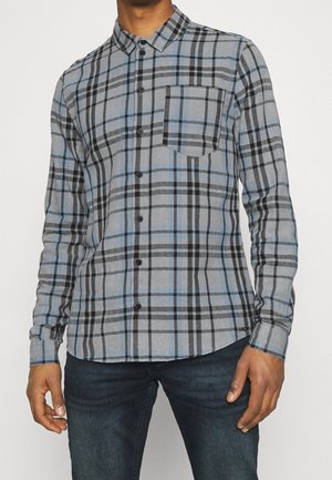 Man wearing blue and gray plaid button-up shirt with black buttons and dark blue jeans against a plain background.