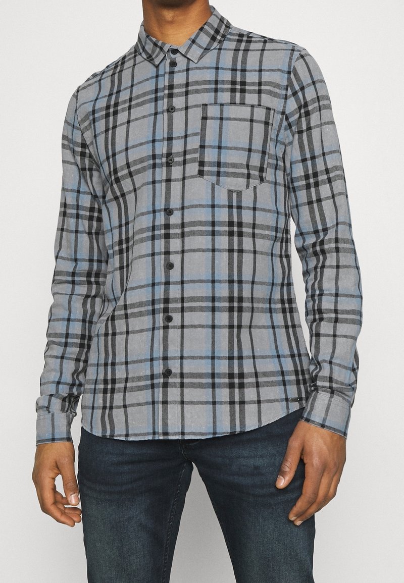 Homme portant une chemise à carreaux bleue et grise avec des boutons noirs et un jean bleu foncé, sur un fond uni.