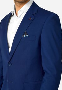 Vīrietis valkā baltu krekla krekliņu un pieguļošu tumši zilu žaketi ar rakstainu kabatas lakatiņu un oranžām šuvēm uz atloka.