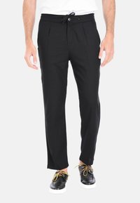 Pantaloni neri a pieghe con vita elastica e cordoncino, caratterizzati da una vestibilità comoda e una texture di tessuto liscia. Indossati con scarpe nere e lacci in contrasto.