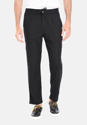 Pantaloni neri a pieghe con vita elastica e cordoncino, caratterizzati da una vestibilità comoda e una texture di tessuto liscia. Indossati con scarpe nere e lacci in contrasto.