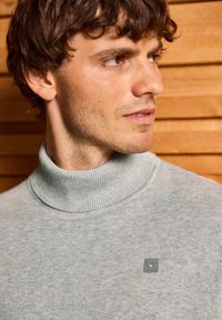 Pull col roulé gris clair en tissu doux, avec un col côtelé et une petite étiquette de marque sur la poitrine.