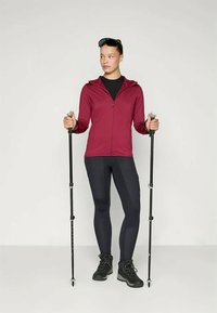 Mladá žena v červené bundě a černých legínách držící turistické hole, oblečená v černých botách a se slunečními brýlemi na hlavě, stojící uvnitř.