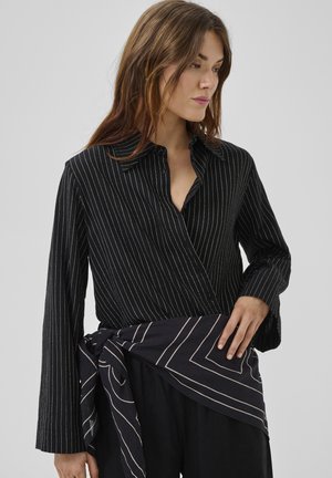 Frau trägt eine schwarze Wickelbluse mit Nadelstreifen, langen Ärmeln und einem dazu passenden Stoffgürtel, der in der Taille gebunden ist.