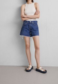 Siniset farkkushortsit, joissa on räjähtänyt helma, yhdistettynä ribbitettyyn kermaiseen topiin ja valkoisiin avonaisiin sandaaleihin, joissa on tummat pohjat.