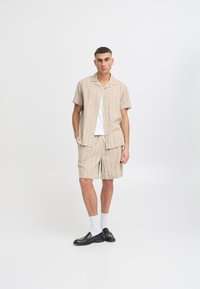 Chemise beige à manches courtes avec de fines rayures verticales, assortie à un short. Le modèle porte des chaussures noires et des chaussettes blanches. Design minimaliste.