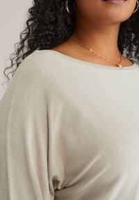 Világos bézs, oversized kötött felső széles dekoltárral és texturált anyaggal. Simító felület jellemzi, és lágyan hullámzik a vállon.