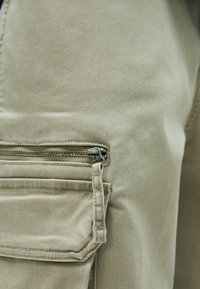Béžové cargo nohavice s bočným zipsovým vreckom a kovovým zipsom. Tkanina má hladkú textúru a odolné švy.
