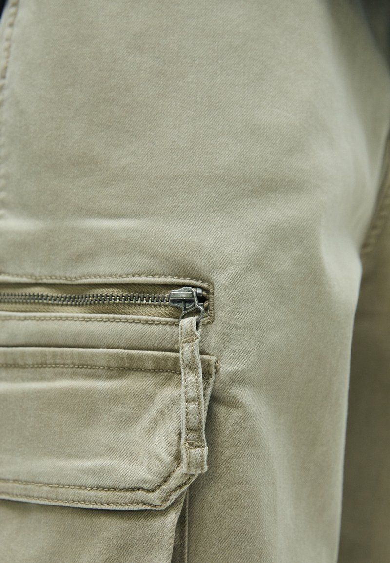 Beige Cargo-Hosen mit einer seitlichen Reißverschlusstasche und einem Metallreißverschluss. Der Stoff hat eine glatte Textur und strapazierfähige Nähte.