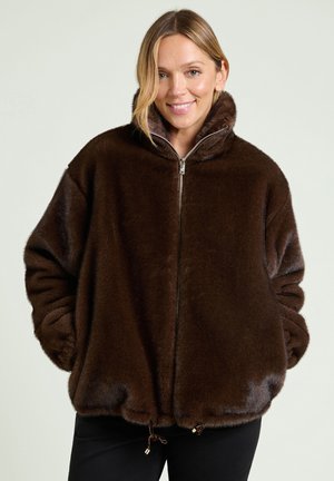 Smiling woman wearing a dark brown faux fur zip-up jacket with hands in pockets against a plain light background.