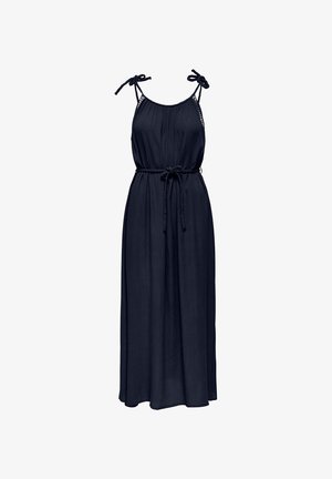 Woman in a long, dark blue sleeveless dress with tied shoulder straps and a thin belt, wearing woven slip-on shoes, looking to the side.