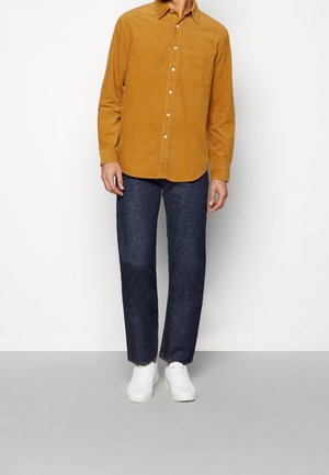 Camisa de pana mostaza con un bolsillo en el pecho, diseño con botones; combinada con vaqueros oscuros y zapatillas blancas. Atuendo sencillo y casual.