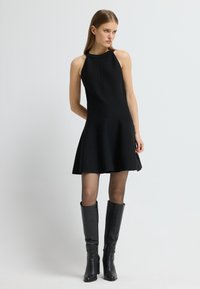 Zwarte mouwloze jurk met een uitlopende rok, gemaakt van een soepele stof. Draag het met hoge zwarte leren laarzen, met een aansluitende silhouet en een zijrits.