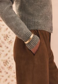 Grauer Strickpullover mit gerippten Bündchen, dazu ein dicker goldener Armreif als Accessoire. Die braunen Wildlederhosen haben eine lockere Passform und Seitentaschen.