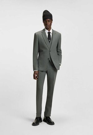 Completo a scacchi grigio con taglio slim, abbinato a una camicia bianca, cravatta nera e berretto di maglia. Scarpe nere completano il look.