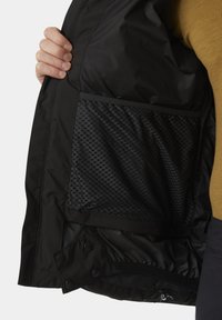 Schwarzes Jackeninnere mit einer Mesh-Tasche, die ein sechseckiges Muster aufweist. Glattes Gewebe mit matter Oberfläche, gesichert mit Druckknöpfen am Saum.