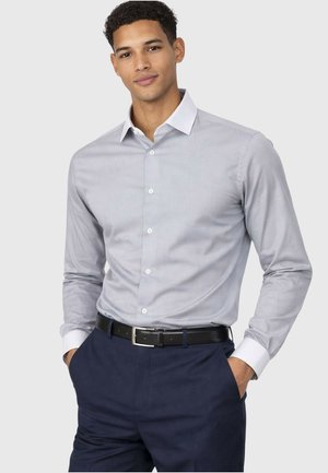 Jeune homme portant une chemise gris clair à boutons avec des poignets blancs, un pantalon bleu marine et une ceinture noire, debout les mains dans les poches.