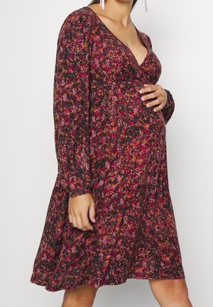 Femme enceinte en robe fluide à manches longues avec motif floral et décolleté en V profond, main posée délicatement sur son ventre arrondi, sur un fond uni.