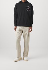 Felpa nera con cappuccio e logo circolare sul petto. Abbinata a pantaloni cargo beige chiaro e sneakers bianche, con tessuto strutturato.