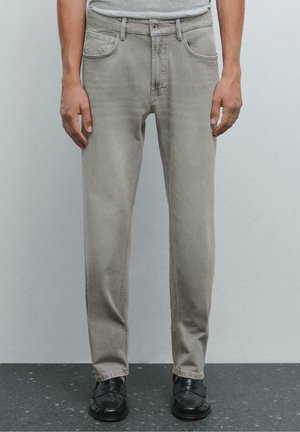Homme portant un jean gris clair droit, une chemise grise rentrée et des mocassins en cuir noirs, debout sur un sol sombre tâcheté.