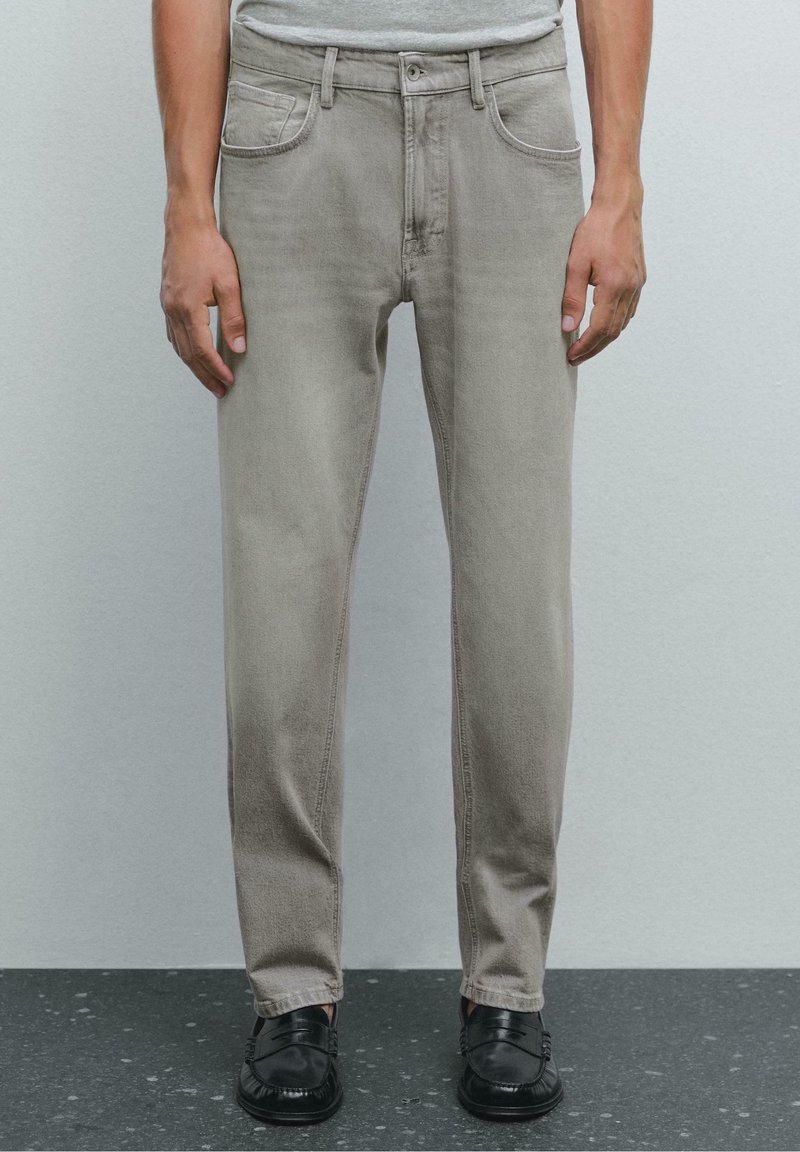 Homme portant un jean gris clair droit, une chemise grise rentrée et des mocassins en cuir noirs, debout sur un sol sombre tâcheté.