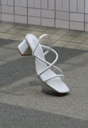Λευκό σανδάλι με λουριά και τακούνι, με τετράγωνη μύτη και χοντρό τακούνι, πάνω σε ασφαλτοστρωμένη επιφάνεια κοντά σε τοιχο με πλακάκια.