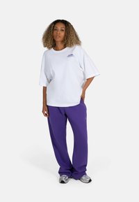 White oversized t-shirt with purple text and logo, paired with loose purple pants. Grey athletic shoes complete the outfit.