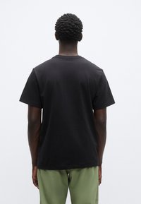 Μαύρο βαμβακερό t-shirt με κλασική εφαρμογή, κοντά μανίκια και στρογγυλή λαιμόκοψη, φορεμένο με χακί παντελόνι. Η πίσω όψη δεν παρουσιάζει γραφικά στοιχεία.