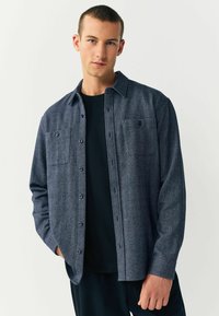 Cameră de cămașă herringbone de culoare albastră închisă, cu guler butonat, două buzunare pe piept și o croială lejeră, purtată peste un tricou negru.