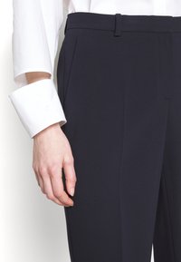 Pantalones de traje azul marino con una textura suave, diseño frontal plano y una pretina delgada. Presentan puños de camisa blanca sobresaliendo, asegurados con botones.