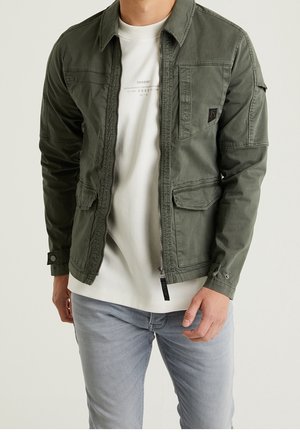 Man met een groen-olijfgroene utility jas met rits open, een wit shirt en lichtgrijze spijkerbroek, staand tegen een effen achtergrond.