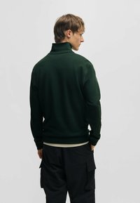 Donkergroen pullover trui met een hoge kraag, voorzien van lange mouwen en een aansluitende tailleband, gecombineerd met zwarte cargobroek.