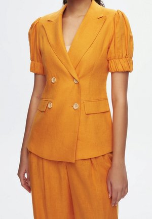 Femme portant un blazer jaune moutarde à double boutonnage avec des manches courtes bouffantes et un pantalon ample assorti sur un fond uni.