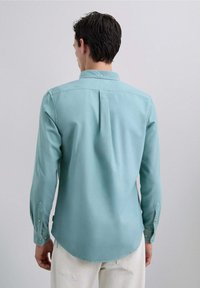 Camisa de botones en algodón azul claro, con mangas largas, cuello puntiagudo y pliegue en la parte posterior. Textura suave con dobladillo curvado.
