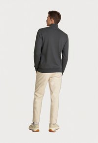 Hombre con suéter de cuello alto gris oscuro, pantalones beige claros y zapatillas beige, de pie con la espalda a la cámara contra un fondo simple.