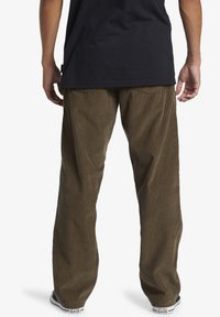 Brown corduroy pants with wide legs, featuring vertical ribbing texture and standard back pockets, worn with a black t-shirt and sneakers.