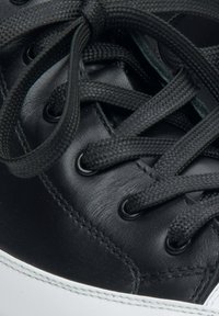 Zwarte leren sneaker met ronde zwarte veters, metalen oogjes en een witte rubberen zool met zichtbare stiksels langs de bovenrand.