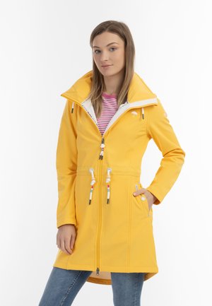 Woman stands with hand in pocket wearing a long yellow raincoat over striped shirt and blue jeans against a white background.