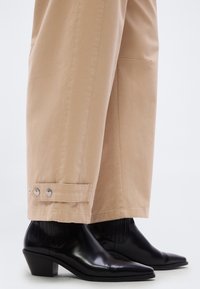 Μαύρα δερμάτινα μποτάκια με μυτερή μύτη, φορεμένα με μπεζ παντελόνια φαρδιάς γραμμής που διαθέτουν λεπτομέρειες με κουμπωμένο λουράκι κοντά στο τελείωμα.