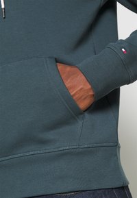 Sudadera de color azul verdoso con un acabado texturizado, que presenta un bolsillo tipo canguro y un acento de cuero marrón. Incluye puños y dobladillo acanalados.