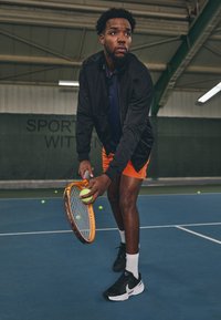 Meestennisist musta jope ja oranžide lühikeste pükstega valmistub siseväljaku serviks, hoides reketit ja palli.